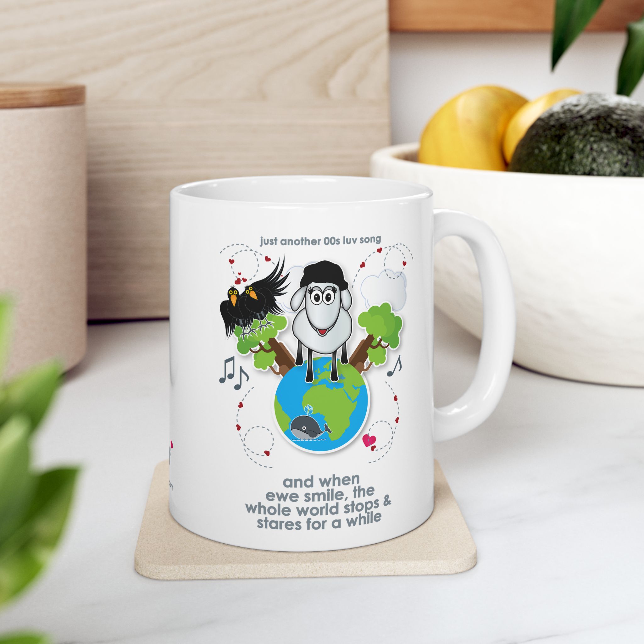 When Ewe Smile Love Song Mug (11oz, 15oz) 6 White ceramic mug printed with a cartoon sheep and some crows on a globe, text reads "and when ewe smile the whole world stops & stares for a while" in kitchen with fruit bowl