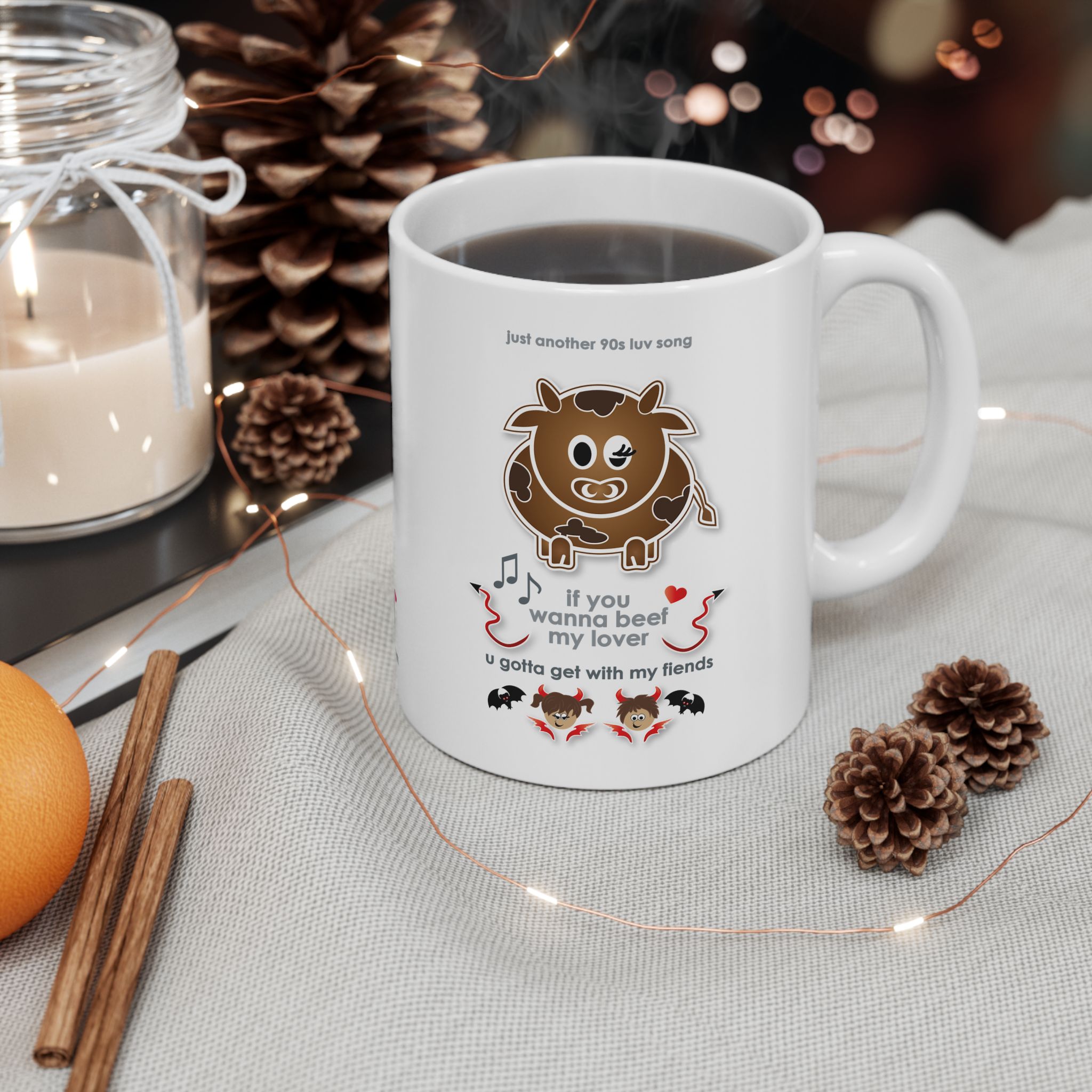 Beef My Lover Love Song Mug (11oz, 15oz) 4 White ceramic Beef my Lover love song mug featuring a cartoon cow, an angel and a fiend, text reads "if you wanna beef my lover, u gotta get with my fiends" on a table with pine cones, cinnamon sticks, a candle, an orange and fairy lights