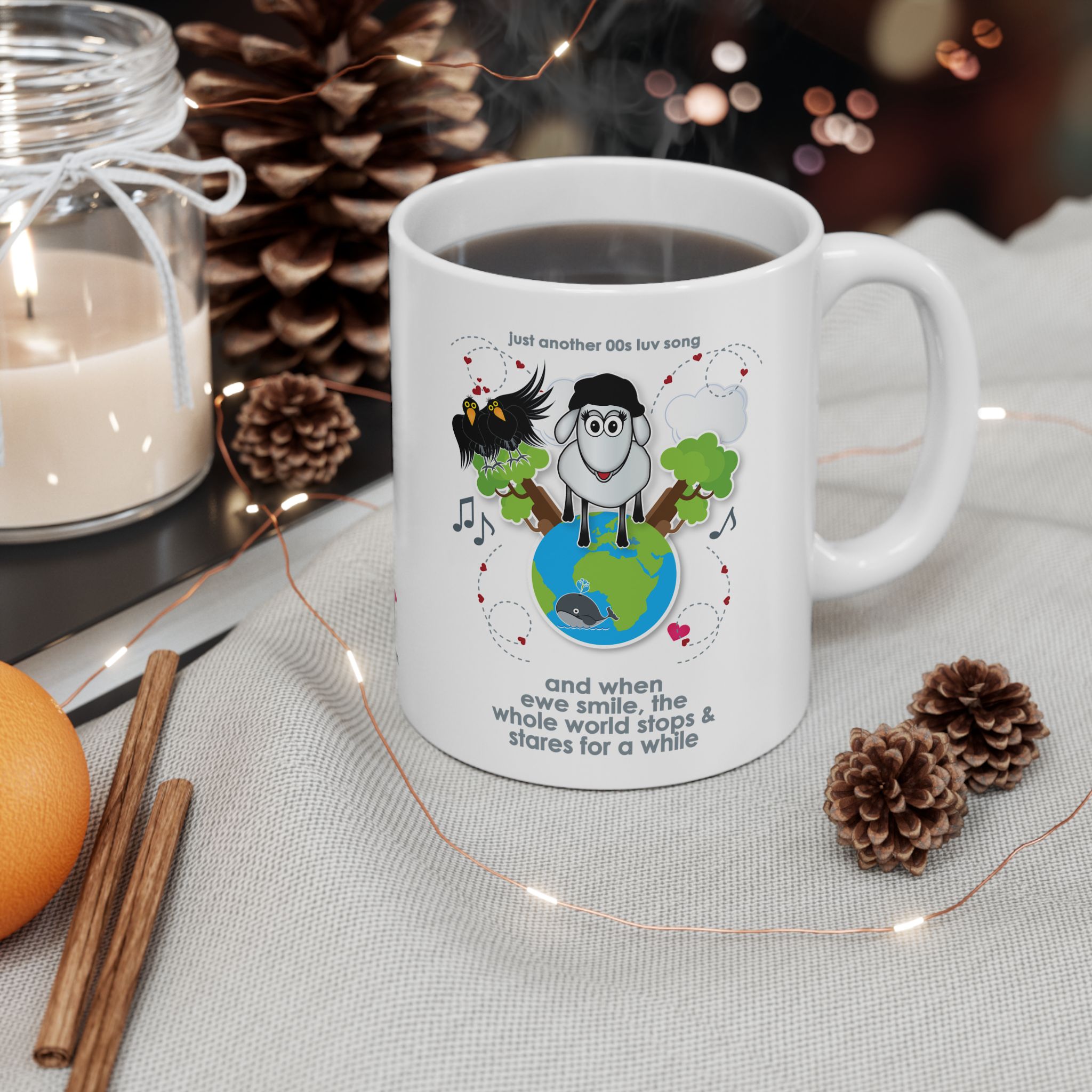 When Ewe Smile Love Song Mug (11oz, 15oz) 4 White ceramic mug printed with a cartoon sheep and some crows on a globe, text reads "and when ewe smile the whole world stops & stares for a while" on a table with pine cones, cinnamon sticks, a candle, an orange and fairy lights